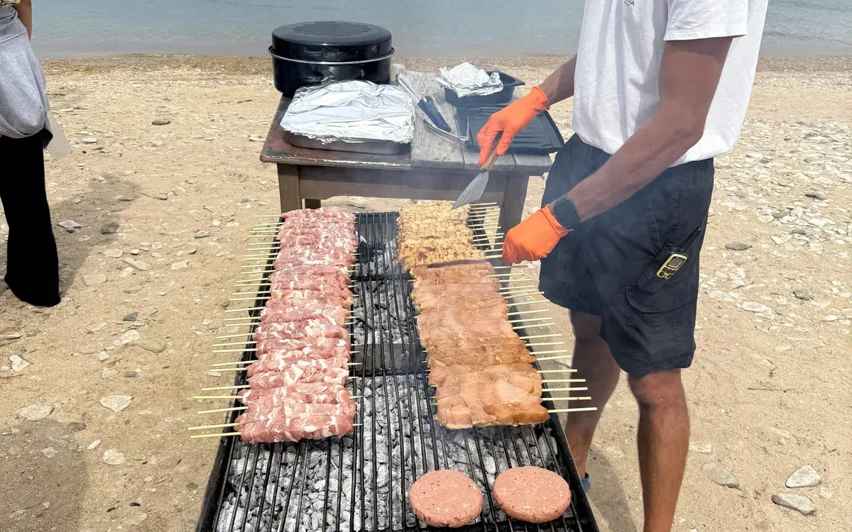 A crew member in orange gloves turning skewers on a charcoal grill on a beach