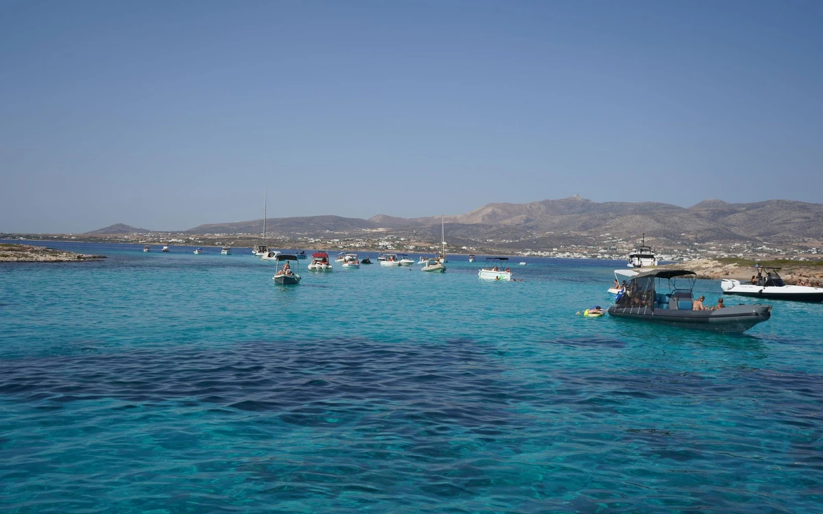 Small boats moored in turquoise shallows, a town and hills on the shore behind