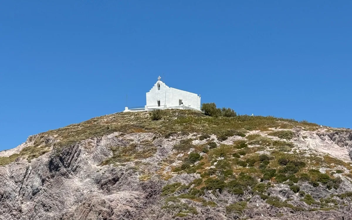 A small white chapel on top of a rocky, scrub-covered islet, seen from the water under a clear blue sky