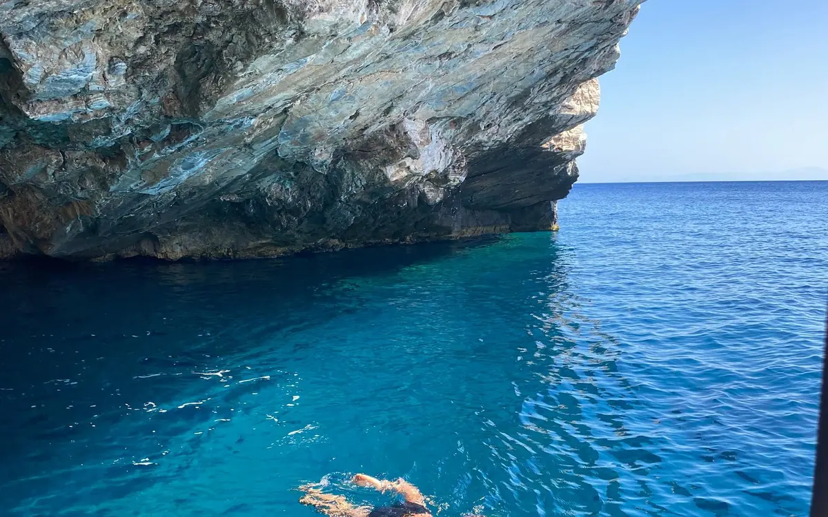 A swimmer in deep blue water below an overhanging rock cliff