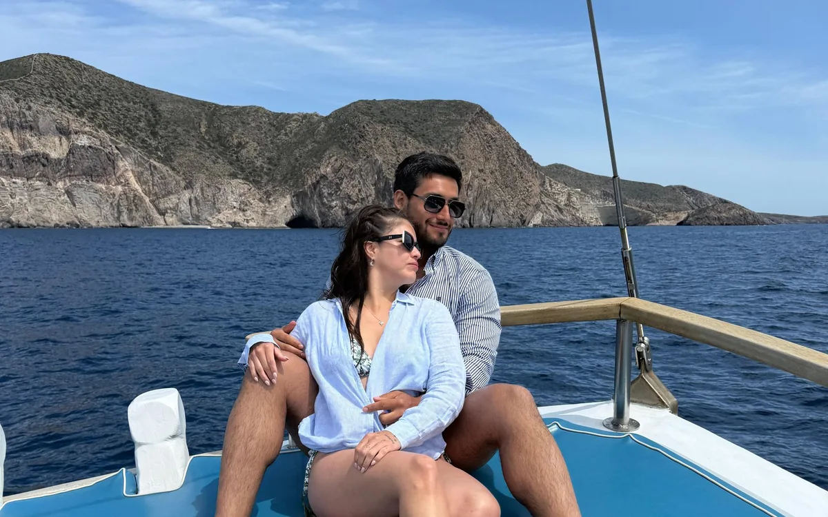 A couple sitting on the bow of a boat with a rocky coastline behind them