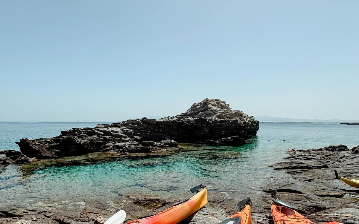 Three orange sea kayaks pulled up on flat dark rocks beside a turquoise rock pool, a rocky islet behind