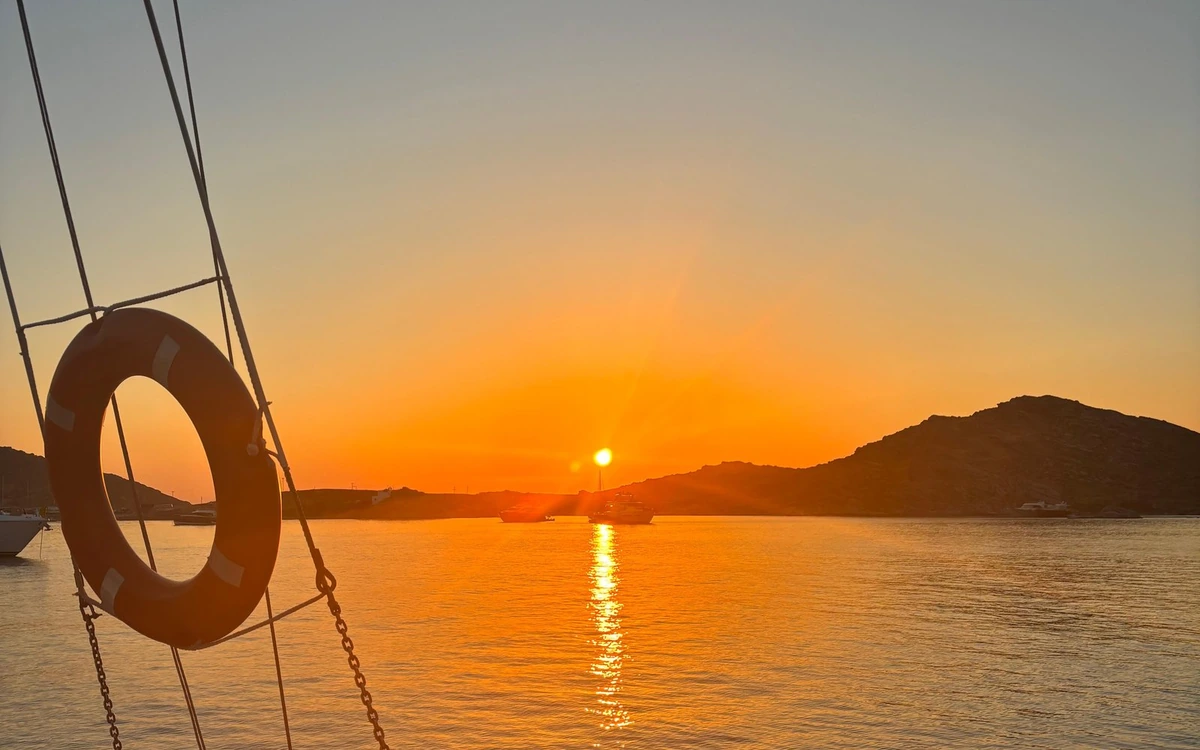 Seen from the deck of a sailing boat: rigging and a lifebuoy in front, the sun setting behind a hill, boats anchored in the bay