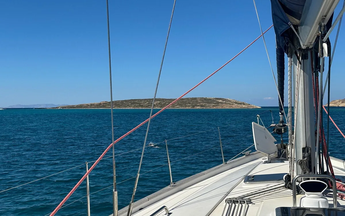 The bow of a sailing boat heading past a low, dry island across deep blue water